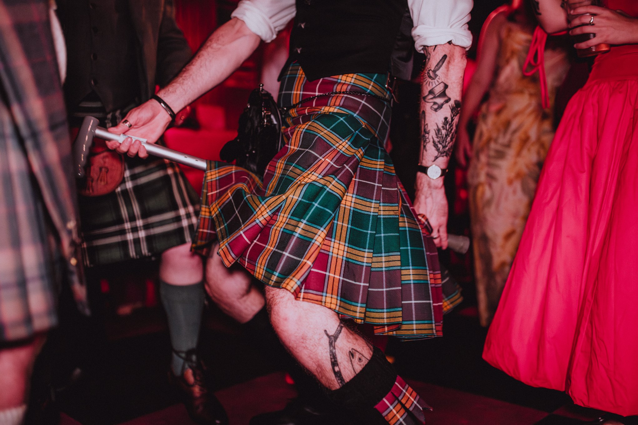 Close-up of wedding guests dancing in kilts during an energetic evening reception, tartan fabrics in motion under warm red dance floor lighting, relax natural wedding photographer Glasgow Scotland