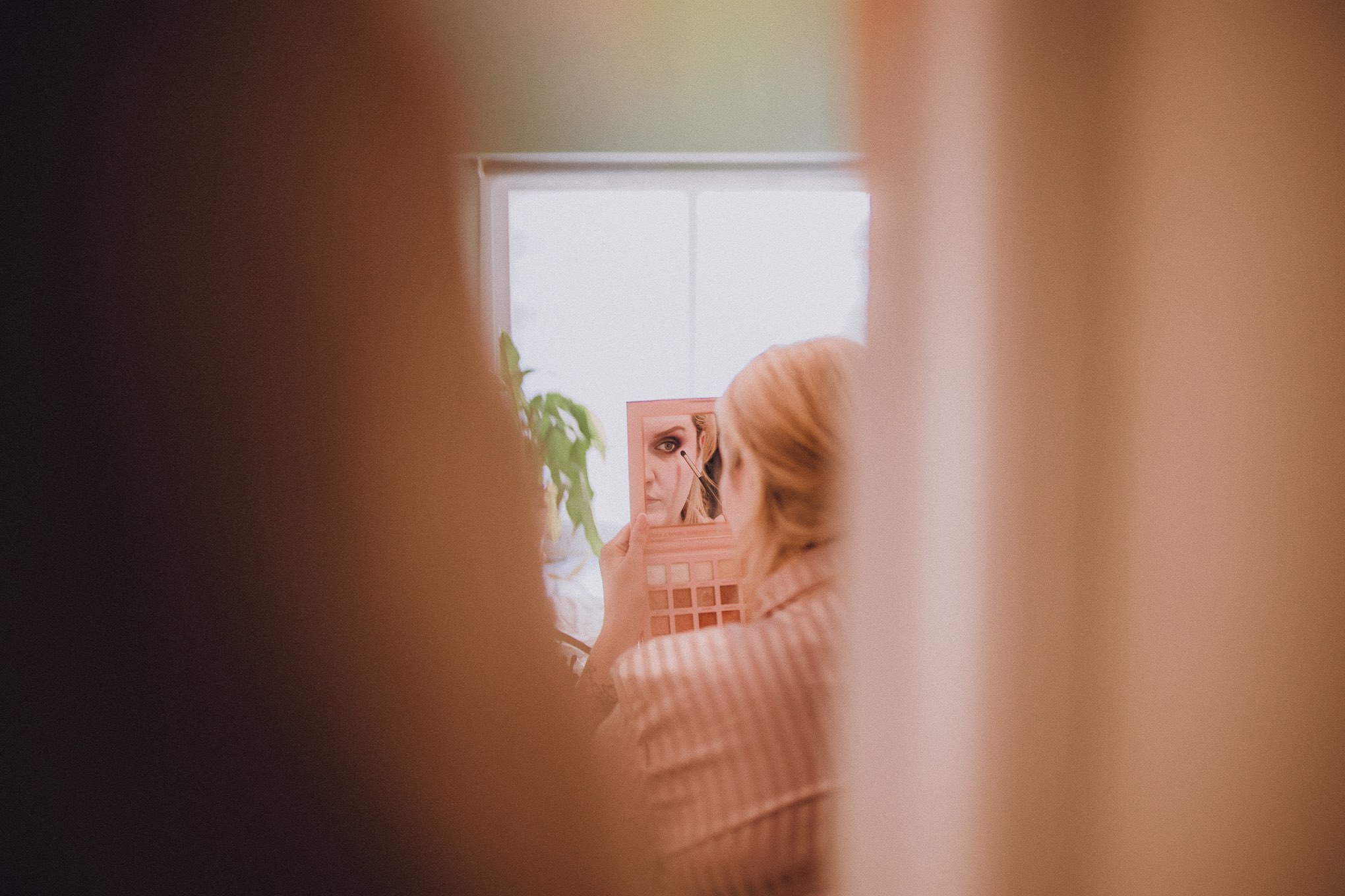 Bride getting ready during morning preparations, seen through a soft foreground frame while applying makeup in natural window light, relax natural wedding photographer Glasgow Scotland