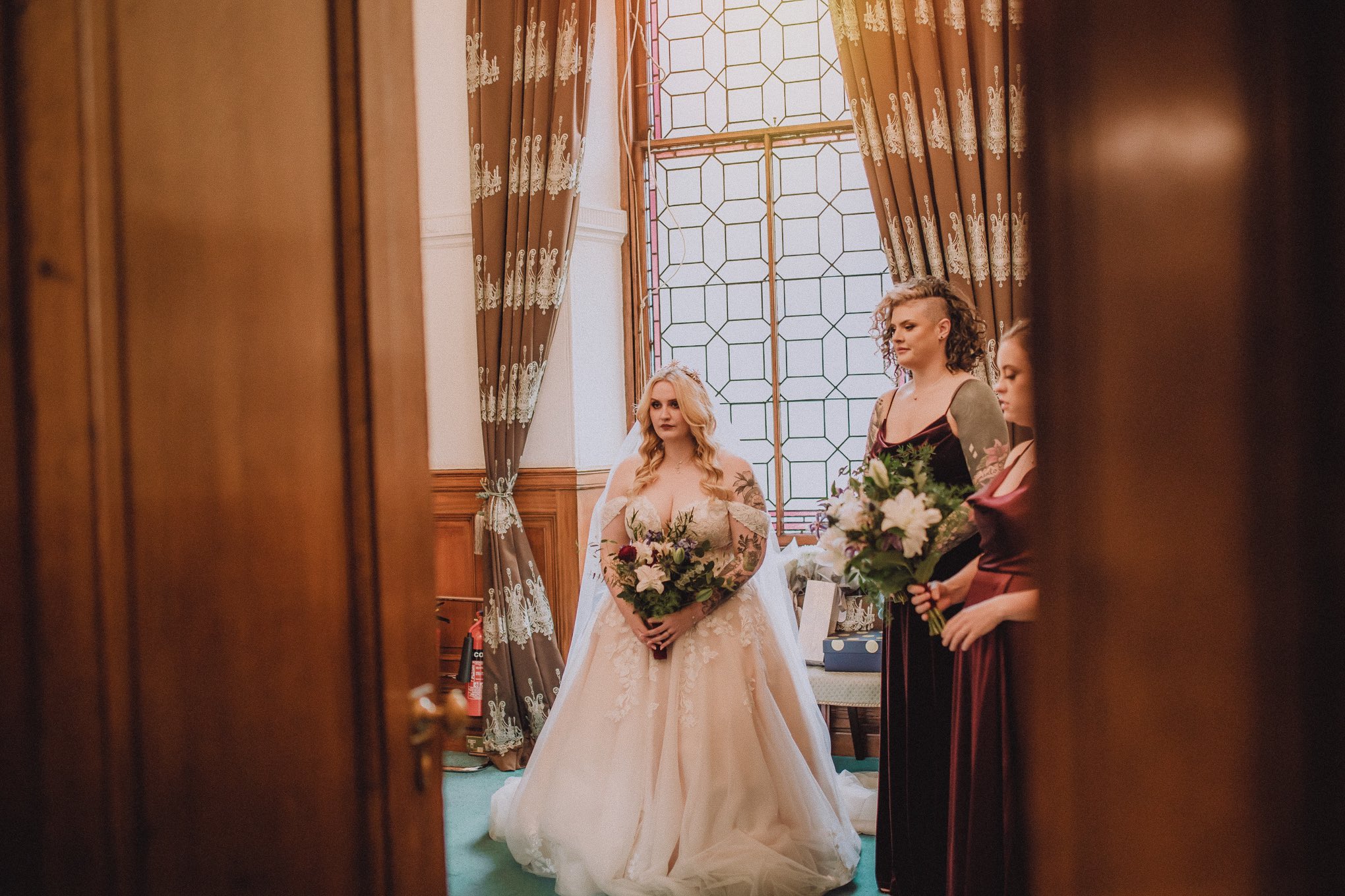 Bride standing with her bridesmaids moments before the ceremony, seen through a doorway with soft light and calm anticipation in the room, relax natural wedding photographer Glasgow Scotland