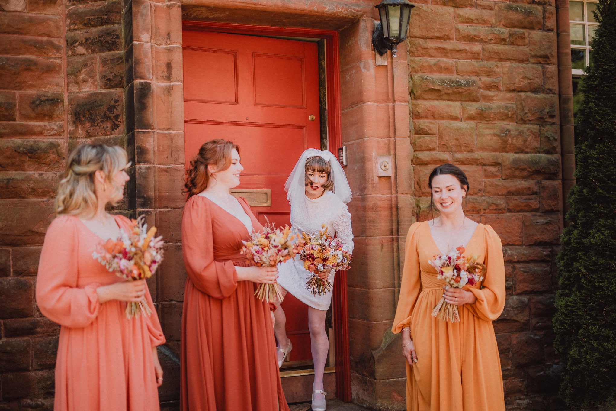 Bride and bridesmaids gathered outside a stone building with a bright red door, warm candid moment filled with laughter and colourful dresses, relax natural wedding photographer Glasgow Scotland