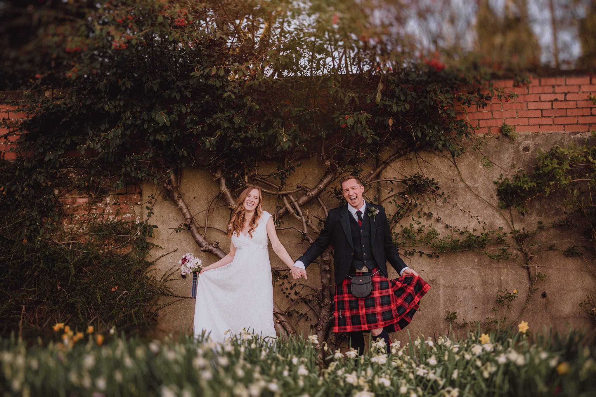Newly married couple holding hands and laughing together outdoors, bride lifting her dress and groom wearing a tartan kilt in a relaxed, joyful moment, relax natural wedding photographer Glasgow Scotland