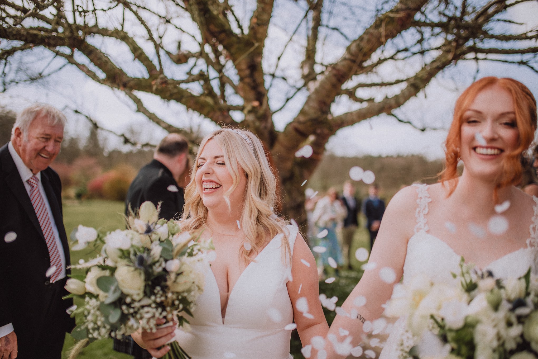 Close-up candid of two brides laughing together during their outdoor wedding ceremony, petals floating through the air with soft natural light, relax natural wedding photographer Glasgow Scotland