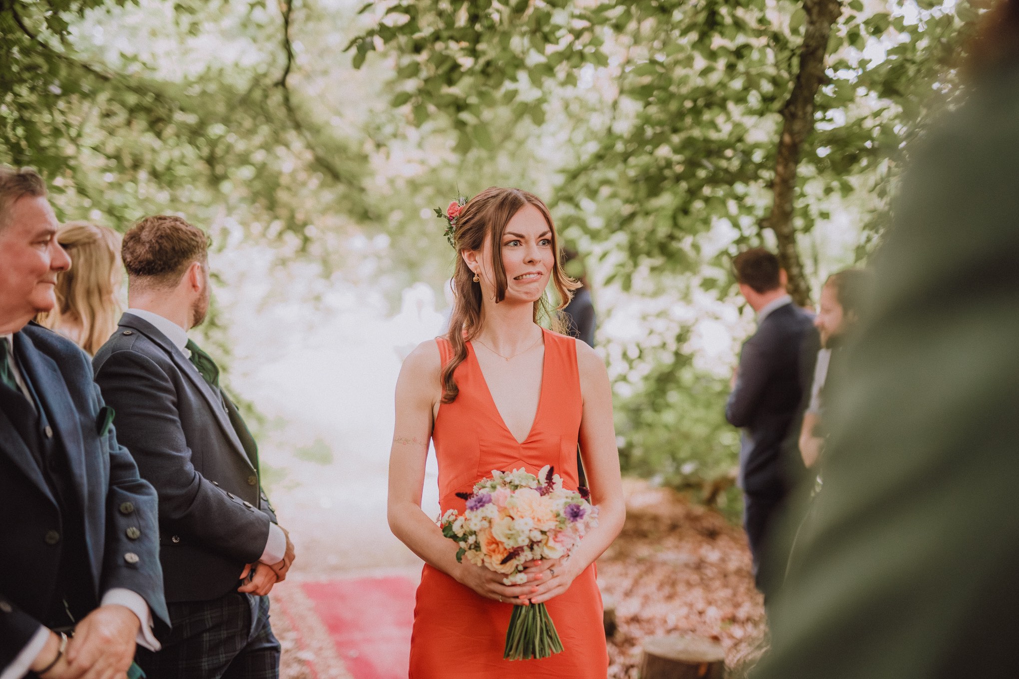 Wedding guest in a burnt-orange dress holding a bouquet during an outdoor woodland ceremony, captured mid-reaction with natural emotion and soft light, relax natural wedding photographer Glasgow Scotland
