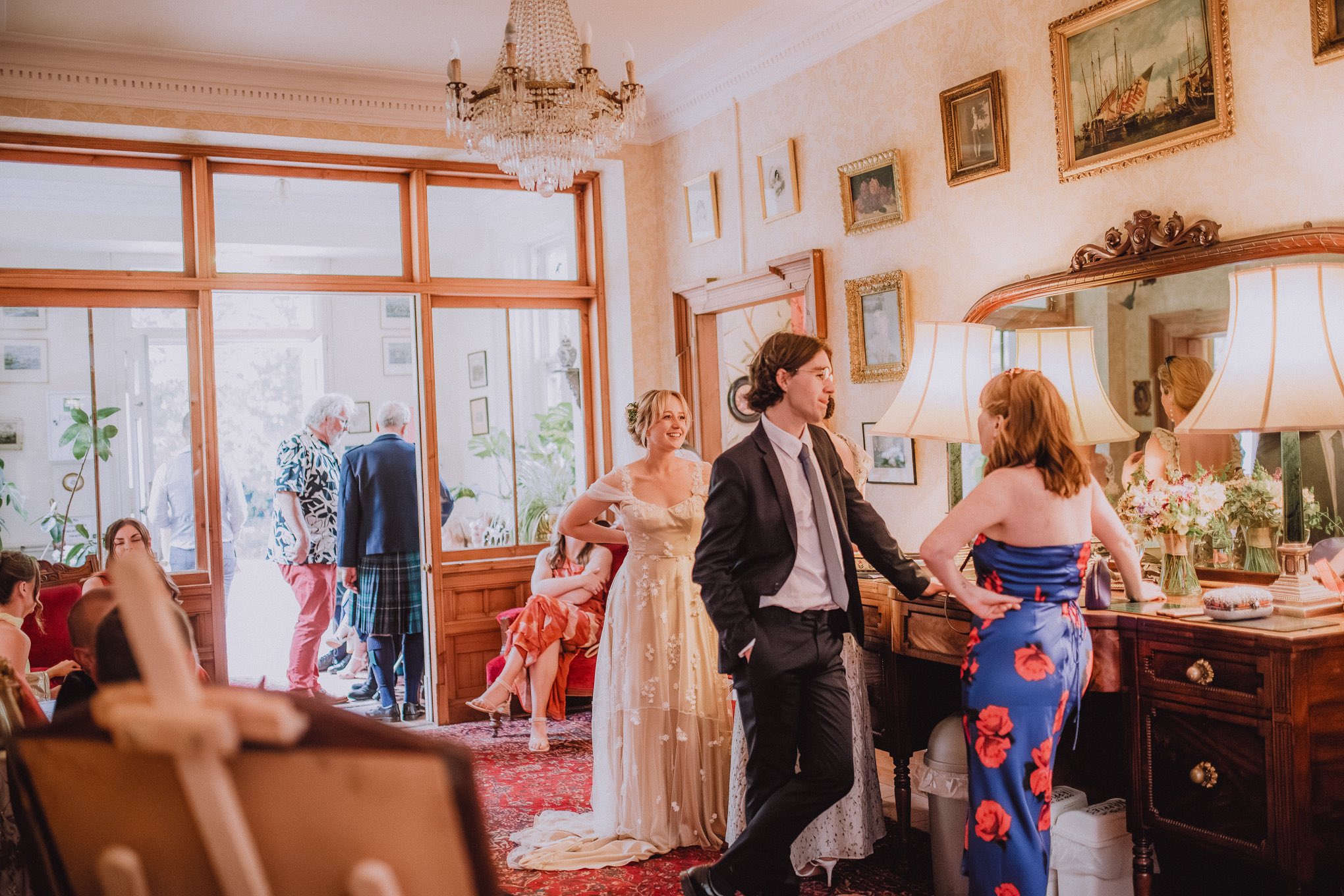Wedding guests chatting and laughing together inside a warm, elegant Scottish venue as the bride smiles in the background, capturing a calm pre-ceremony moment with friends and family, relaxed natural waiting photography Scotland