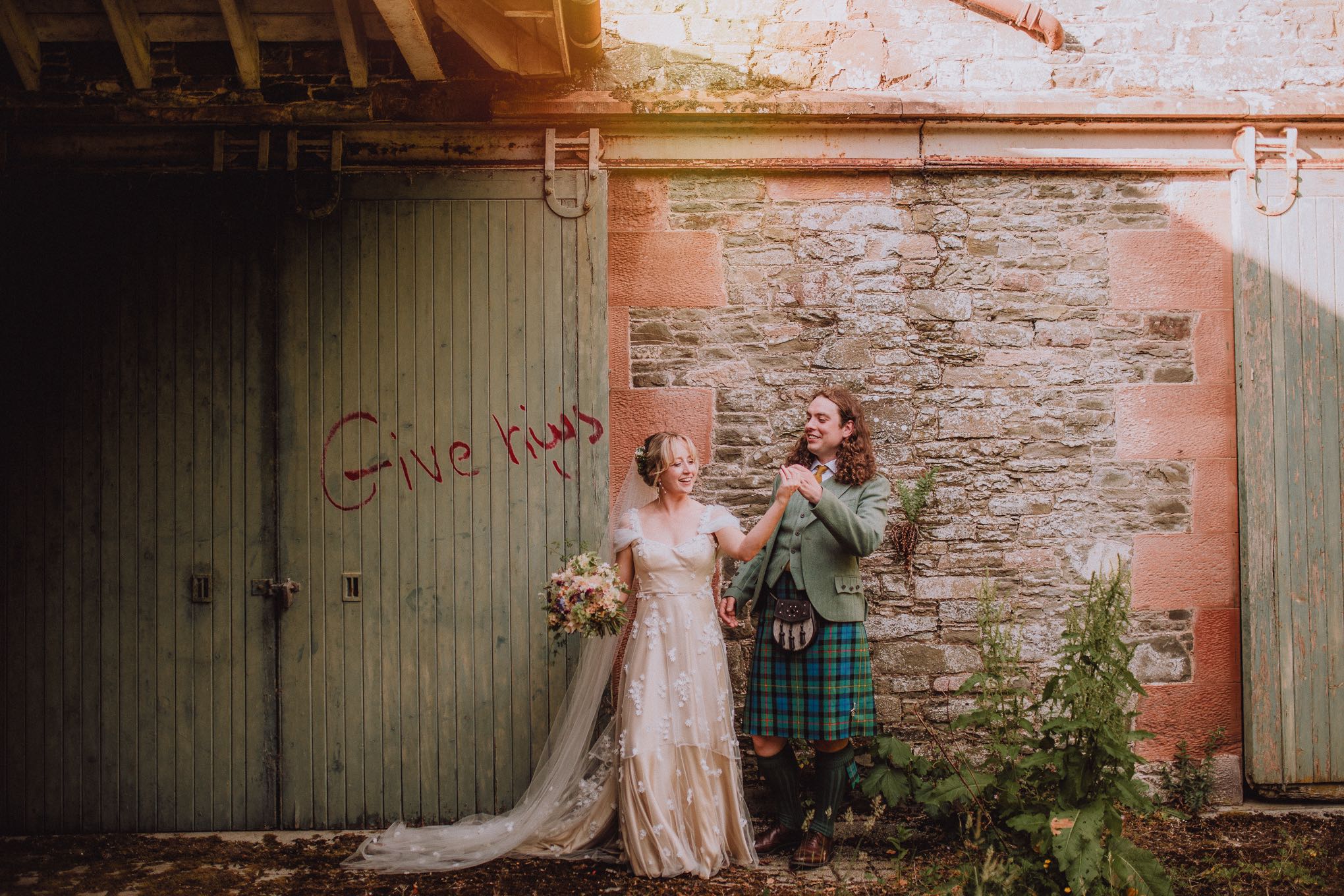 Bride and groom laughing together outside a rustic Scottish stone building beside weathered green barn doors with graffiti, photographed in a relaxed, documentary style by a wedding photographer Scotland