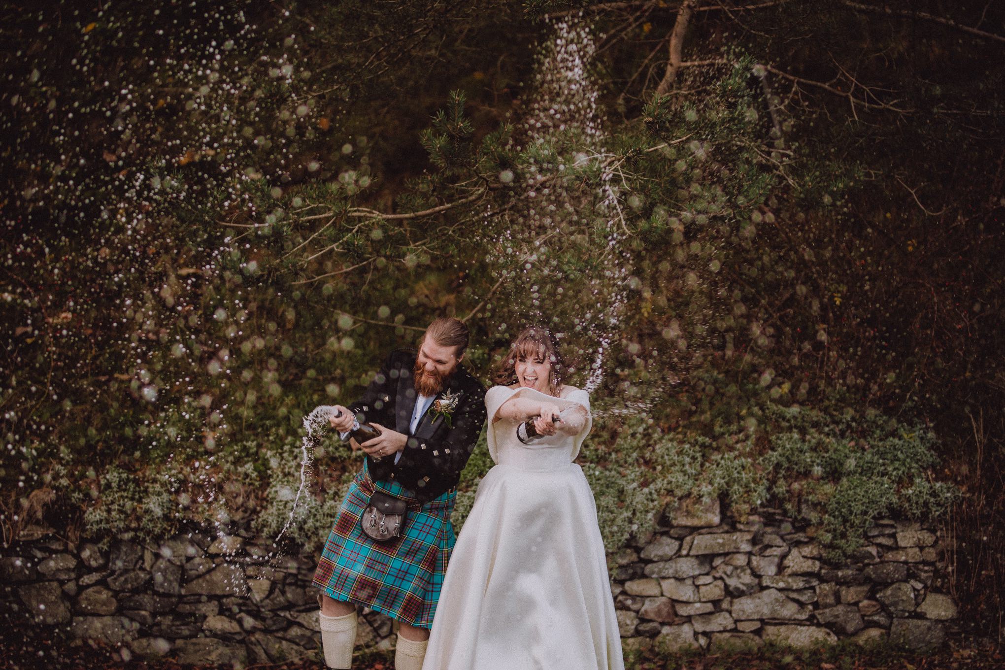 Bride and groom joyfully spraying champagne outdoors against a woodland backdrop in Scotland, captured mid-action in a relaxed documentary style by a wedding photographer Scotland