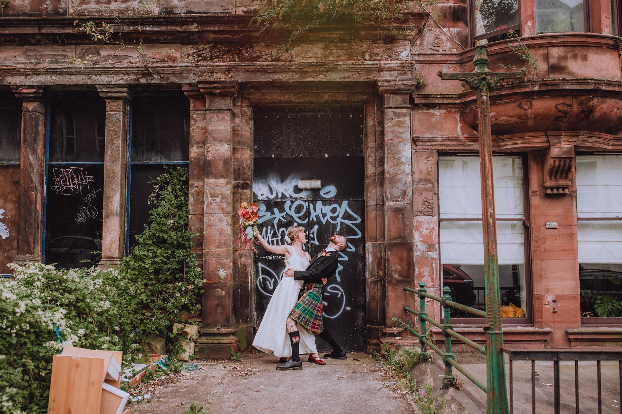 Playful newlywed couple posing outside a weathered sandstone building with graffiti, alternative wedding portrait blending romance and city texture, relax natural wedding photographer Glasgow Scotland